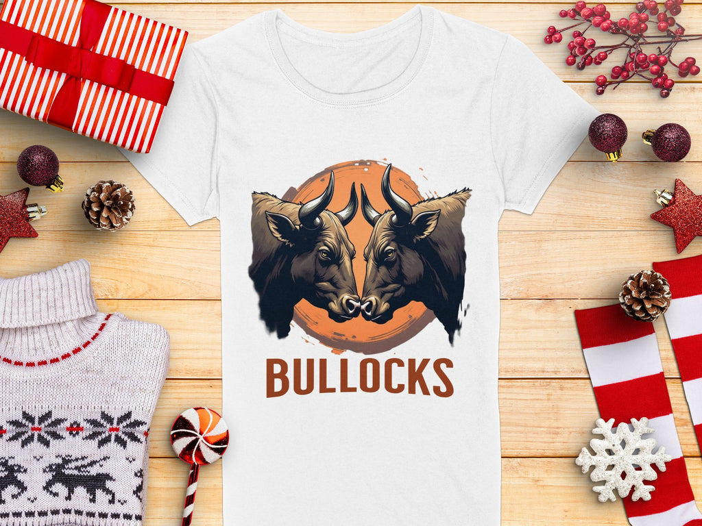 A Garment Graphics white t-shirt features bold graphics with two bulls facing each other above the word BULLOCKS, displayed on wood with wrapped gifts, pinecones, and decorations.
