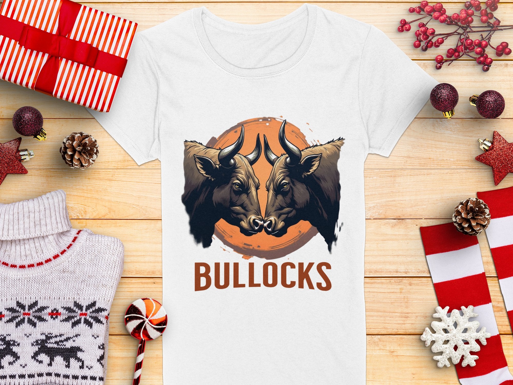 A Garment Graphics white t-shirt features bold graphics with two bulls facing each other above the word BULLOCKS, displayed on wood with wrapped gifts, pinecones, and decorations.