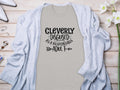 A classic light gray T-shirt by Garment Graphics features Cleverly Disguised As A Responsible Adult in a playful font. Ideal for casual outings, it’s styled on wood with a light blue cardigan, white shoes, and delicate white flowers.