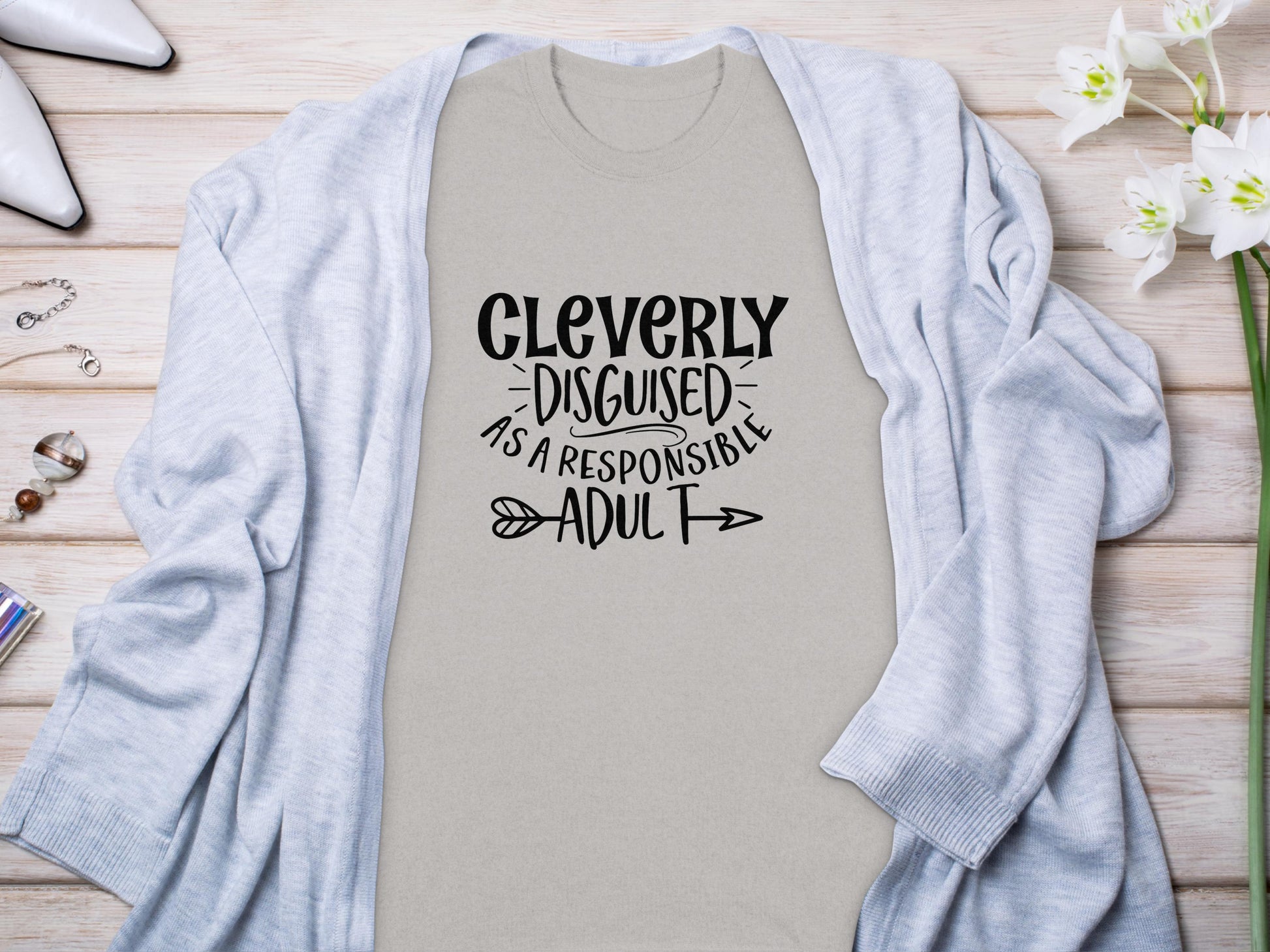 A classic light gray T-shirt by Garment Graphics features Cleverly Disguised As A Responsible Adult in a playful font. Ideal for casual outings, it’s styled on wood with a light blue cardigan, white shoes, and delicate white flowers.