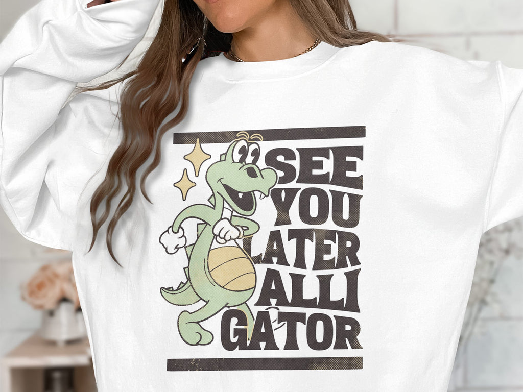 A Garment Graphics sweatshirt features a cartoon alligator with a basketball and the bold phrase SEE YOU LATER ALLIGATOR on its white fabric, with the wearers face not visible.