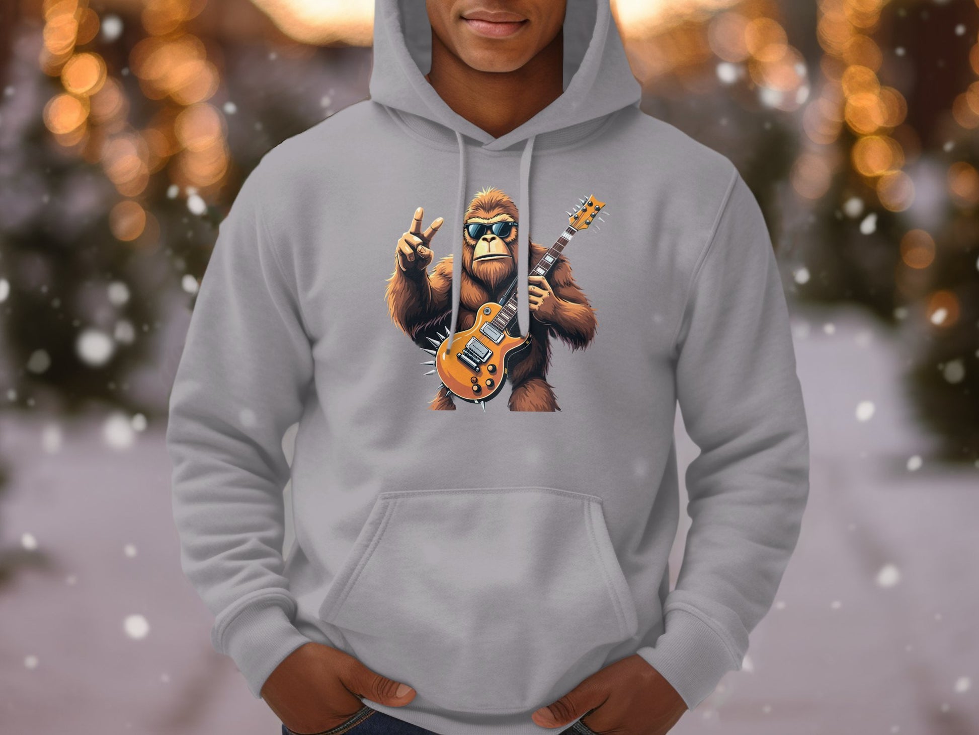 A person in a gray Garment Graphics hoodie features a guitar-playing Bigfoot giving a peace sign. Ideal for animal and music enthusiasts, it includes blurred winter lights and falling snow, enhancing the charm of the Guitar Playing Gorilla Hoodies collection.