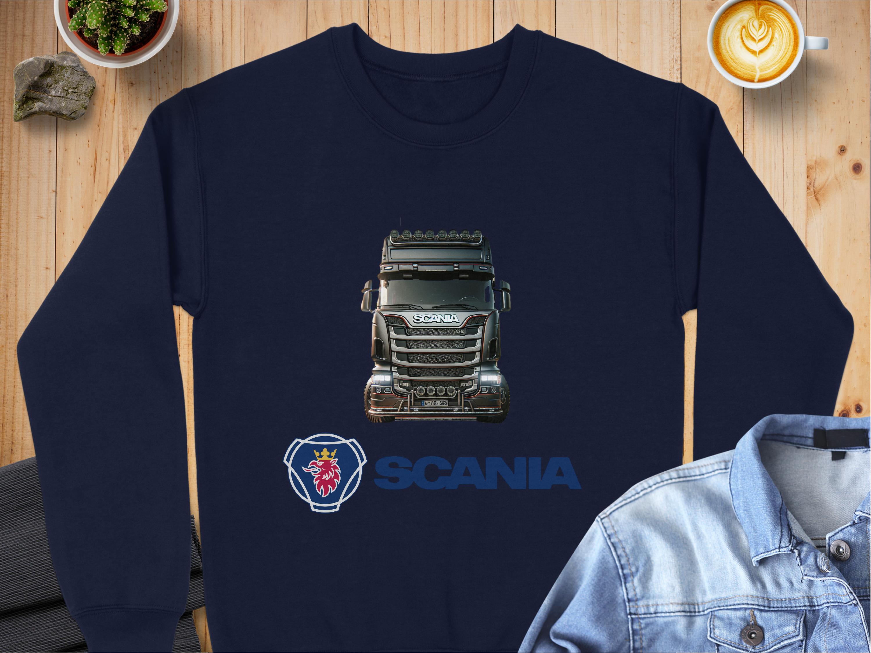 A navy blue graphic crewneck sweatshirt from Garment Graphics showcases a Scania truck image, with the logo and SCANIA in bold blue beneath it. Ideal for truck fans, its styled with a denim jacket, coffee, and potted plant on a wooden surface.