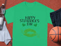 A Garment Graphics T-shirt featuring Happy St. Patricks Day with green lips and clovers sits beside black shorts, a basketball, a water bottle, and a towel.