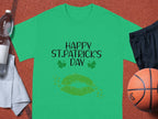 A Garment Graphics T-shirt featuring Happy St. Patricks Day with green lips and clovers sits beside black shorts, a basketball, a water bottle, and a towel.