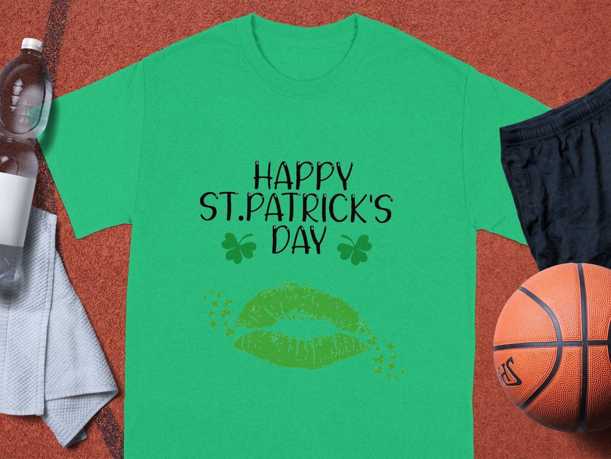 A Garment Graphics T-shirt featuring Happy St. Patricks Day with green lips and clovers sits beside black shorts, a basketball, a water bottle, and a towel.