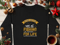 A black Garment Graphics sweatshirt featuring Father and Son: Fishing Partners for Life with a fish and rod design is nestled among festive decorations, like pine branches, ornaments, and a cozy mug of coffee, celebrating the father-son bond.