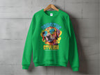 On a concrete wall, a green sweatshirt with a vibrant dog graphic in sunglasses is displayed. Above it reads Pawsitive Stylish, with Create, Play, Dance, Walk below—ideal for dog lovers seeking trendy Dog Graphic Sweatshirts by Garment Graphics.