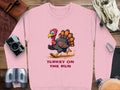 Garment Graphics pink sweatshirt features a cartoon turkey and Turkey on the Run text. Unisex sizes. It is displayed flat on wood, surrounded by a camera, binoculars, leather strap, and beige pouch.