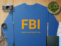 Blue graphic print sweatshirt from Garment Graphics featuring bold yellow FBI: Female Boob Inspector text. Scene includes a camera, lens, bag, notebook, fruit bowl, and map on wood. Designed in unisex sizing for everyone.