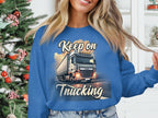 A person in a blue Garment Graphics graphic sweatshirt with a truck and Keep On Trucking stands by a decorated Christmas tree, smiling widely while playing with their hair. The unisex sizing ensures both comfort and style for the festive season.