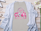 Garment Graphics offers a light beige Just be Cool Rock and Roll T-shirt, featuring pink and white lettering with headphone graphics. Styled under a light gray cardigan, it rests on wood, adorned with jewelry, white flowers, and high heels—a perfect pick for music lovers.