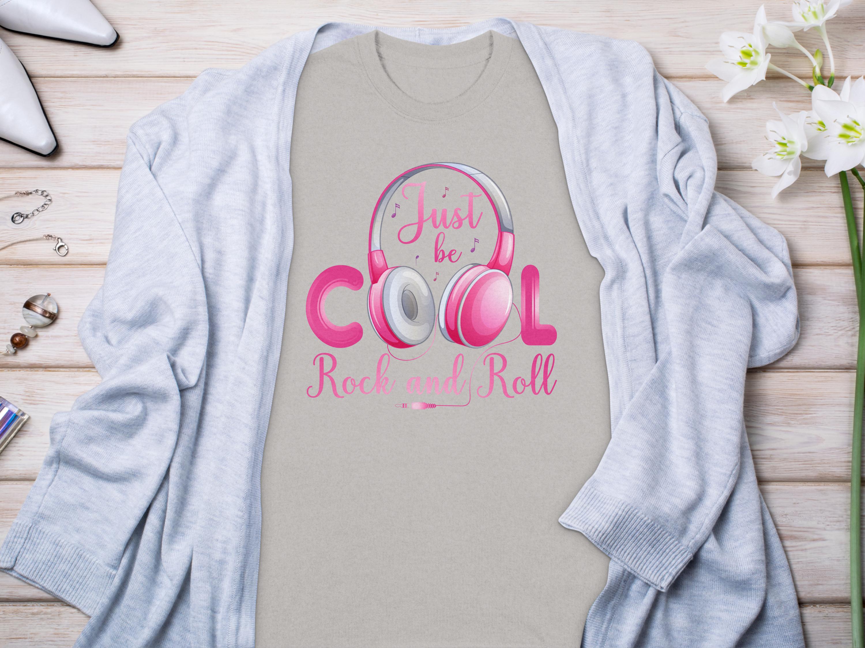 Garment Graphics offers a light beige Just be Cool Rock and Roll T-shirt, featuring pink and white lettering with headphone graphics. Styled under a light gray cardigan, it rests on wood, adorned with jewelry, white flowers, and high heels—a perfect pick for music lovers.
