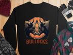 Garment Graphics presents a loose-fit black sweatshirt with an eye-catching bullocks graphic: two illustrated bulls with large horns against an orange circle and BULLOCKS boldly printed below, displayed on wood with cozy winter accessories nearby.