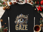 The Garment Graphics black unisex sweatshirt features an ornate Gaze eye graphic, surrounded by festive elements like pine branches, red and gold ornaments, pinecones, stars, and a cup of tea on a wooden surface.