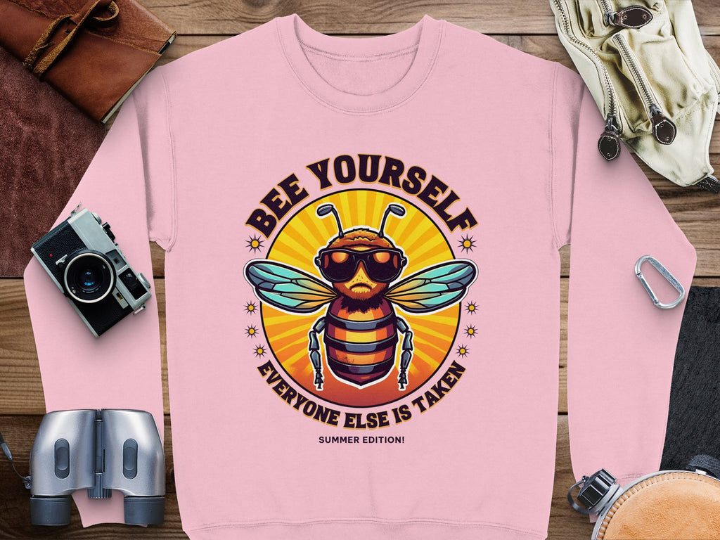 Garment Graphics presents a pink sweatshirt with a cartoon bee in sunglasses and the motto BEE YOURSELF, EVERYONE ELSE IS TAKEN. This unisex summer edition features a sunburst background and playful icons like a camera, compass, and binoculars to inspire adventure.
