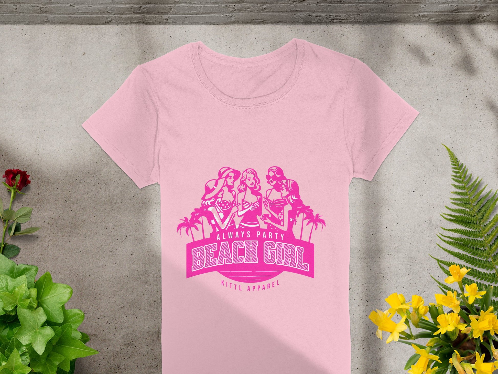 A pink t-shirt from Garment Graphics Beach Girl T-Shirts collection is showcased on a concrete surface. It features a graphic of three women with palm trees and the text Always Party Beach Girl in bold pink letters, surrounded by green plants and yellow flowers.