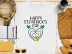 The Garment Graphics festive sweatshirt with Happy St. Patricks Day and green clovers pairs well with a plaid scarf. Also featured are a black pen, notebook, yellow purse, sunglasses, quilted jacket, and striped coffee mug on wood complementing the clover design ensemble.