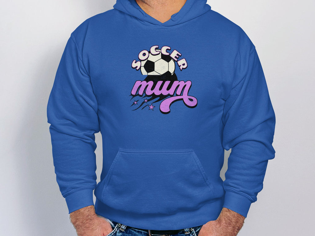 Wearing a cozy blue hoodie by Garment Graphics, a person stands against a plain background. The classic fit features Soccer Mum in bold and an illustration of a soccer ball with stars above it, offering warmth and comfort.