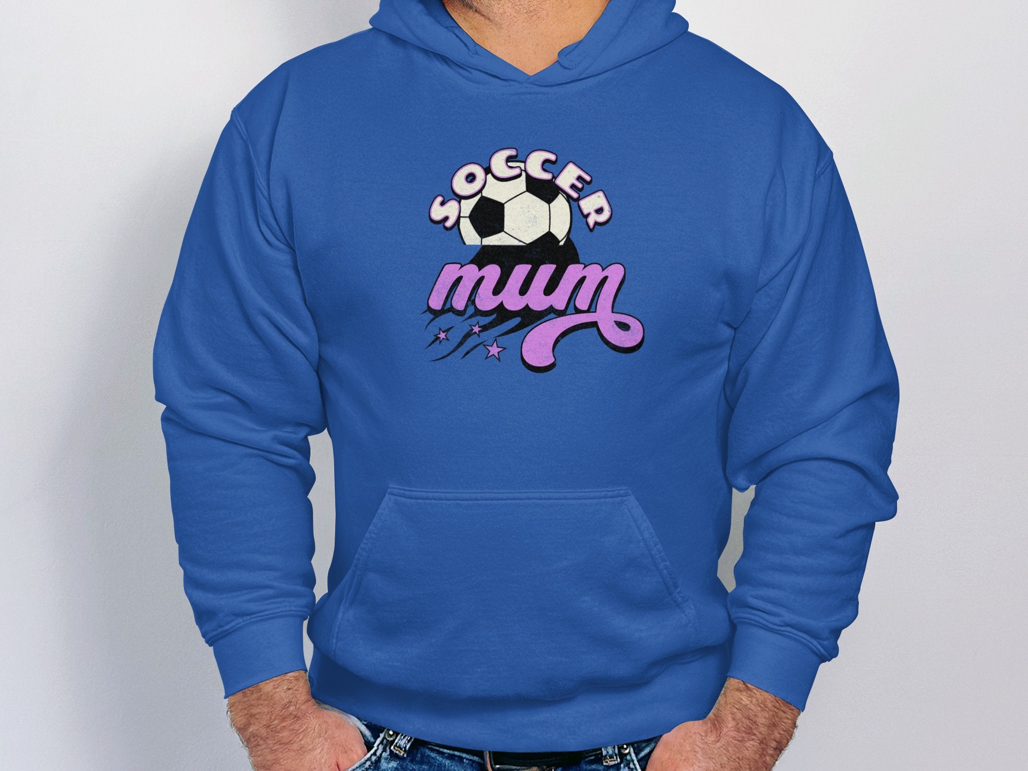 Wearing a cozy blue hoodie by Garment Graphics, a person stands against a plain background. The classic fit features Soccer Mum in bold and an illustration of a soccer ball with stars above it, offering warmth and comfort.