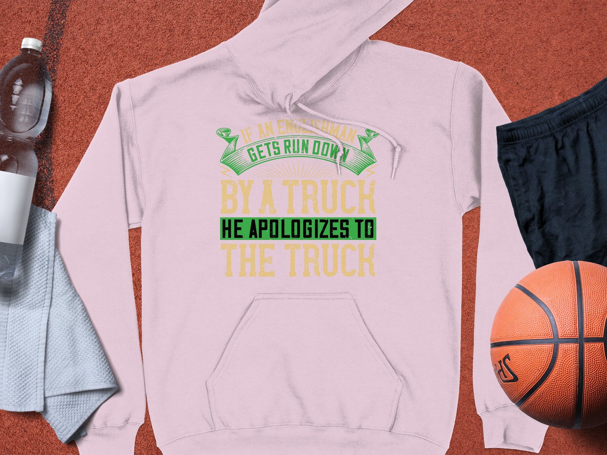 A light pink hoodie by Garment Graphics showcases the witty quote If an Englishman gets run down by a truck, he apologizes to the truck in green and yellow. Its humorously displayed with a water bottle, towel, black shorts, and a basketball on a red surface.