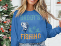 A person in a blue Daddys Fishing Buddy sweatshirt, featuring a fish and boat graphic, poses by a festive Christmas tree. Perfect for fishing fans, the scene includes shelves with white decorations. Brand: Garment Graphics.