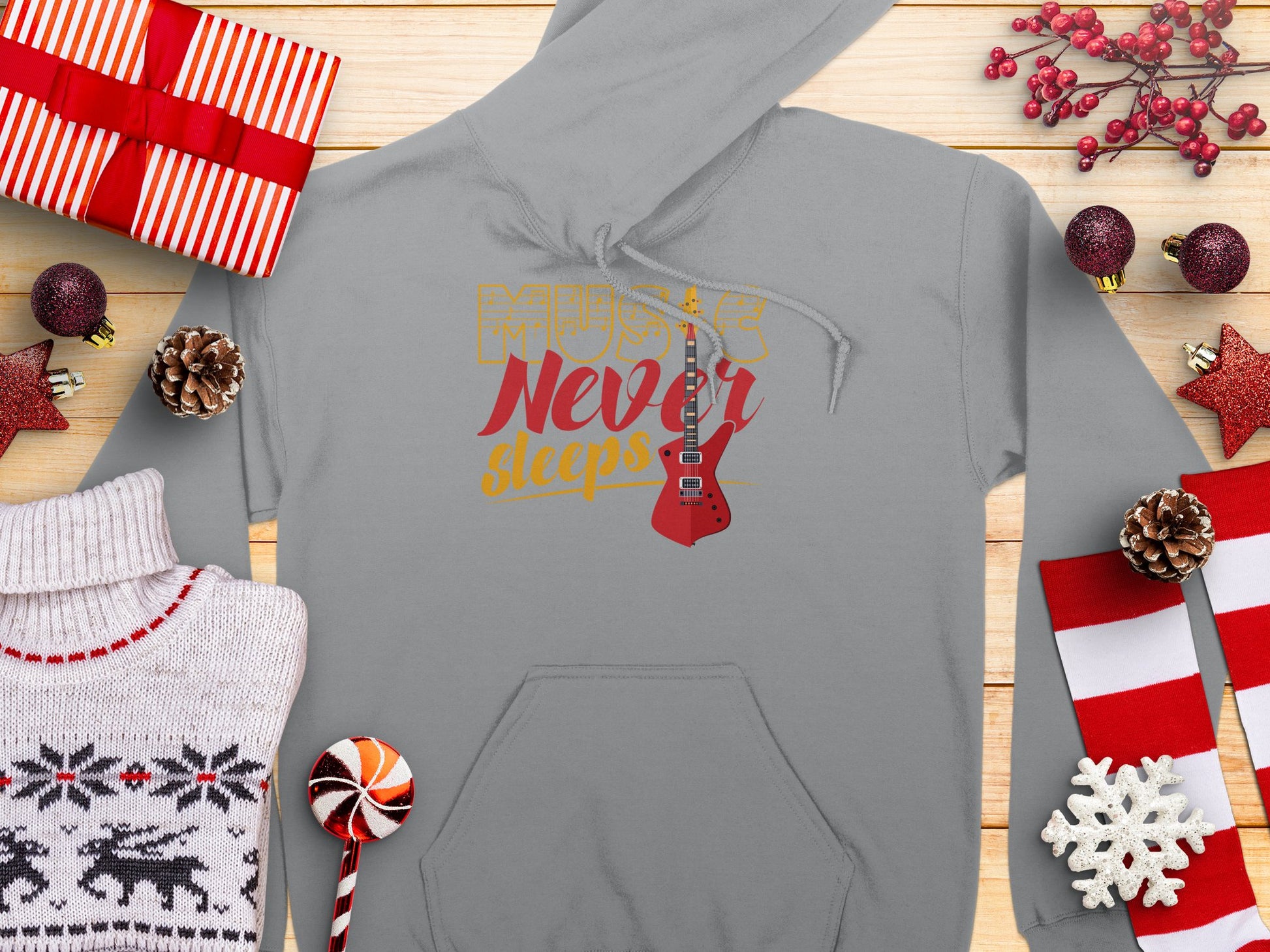 A Garment Graphics gray hoodie featuring Music Never Sleeps with a guitar design lies on a wooden surface surrounded by gifts, a sweater, candy, pine cones, and red decorations. Its perfect for any music lover who appreciates classic fit and style.