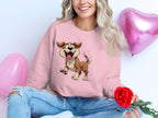 Dressed in a pink Garment Graphics sweatshirt with a cheerful animated dog design, a person sits cross-legged, smiling. They hold a red rose amid two pink heart-shaped balloons, showcasing joyful love for dogs and wearing jeans torn at the knees.