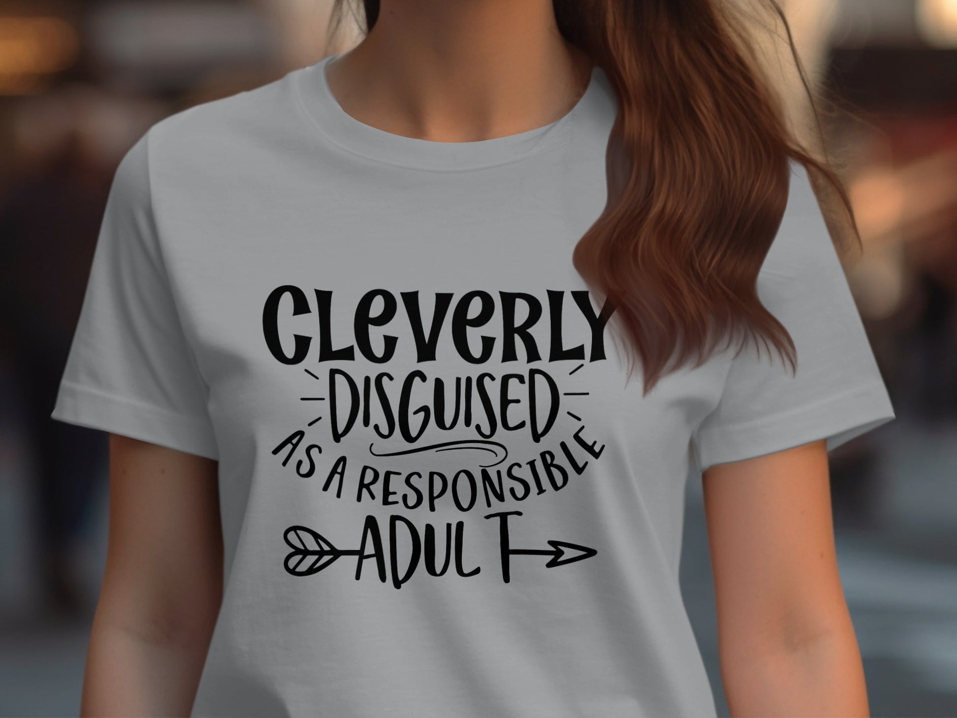 A woman wears a semi-fitted light gray Garment Graphics T-shirt featuring the playful text Cleverly disguised as a responsible adult in bold black letters with an arrow, set against a blurred background that adds depth to her cheeky outfit.