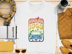 A white T-shirt from Garment Graphics featuring a retro Woodstock Festival 1969 peace sign and bird design lies on a wooden table. Complemented by yellow sunglasses, quilted purse, checked scarf, notebook and pen, mustard jacket, and mug, it exudes vintage vibes.