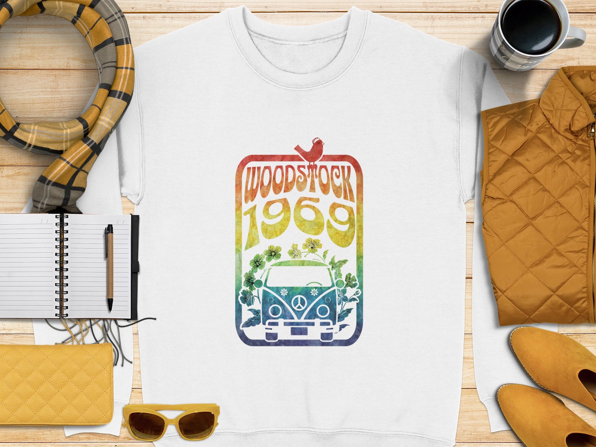 A white T-shirt from Garment Graphics featuring a retro Woodstock Festival 1969 peace sign and bird design lies on a wooden table. Complemented by yellow sunglasses, quilted purse, checked scarf, notebook and pen, mustard jacket, and mug, it exudes vintage vibes.