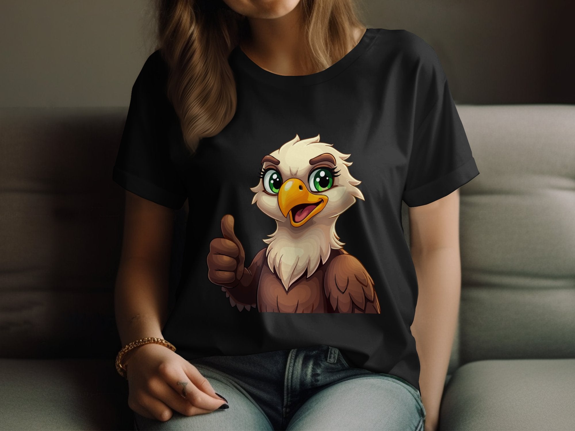 A person on a couch models a Garment Graphics eagle T-shirt, featuring a cartoon eagle with large green eyes, brown feathers, and giving a thumbs-up. The design suits animal lovers while keeping the persons face out of view.