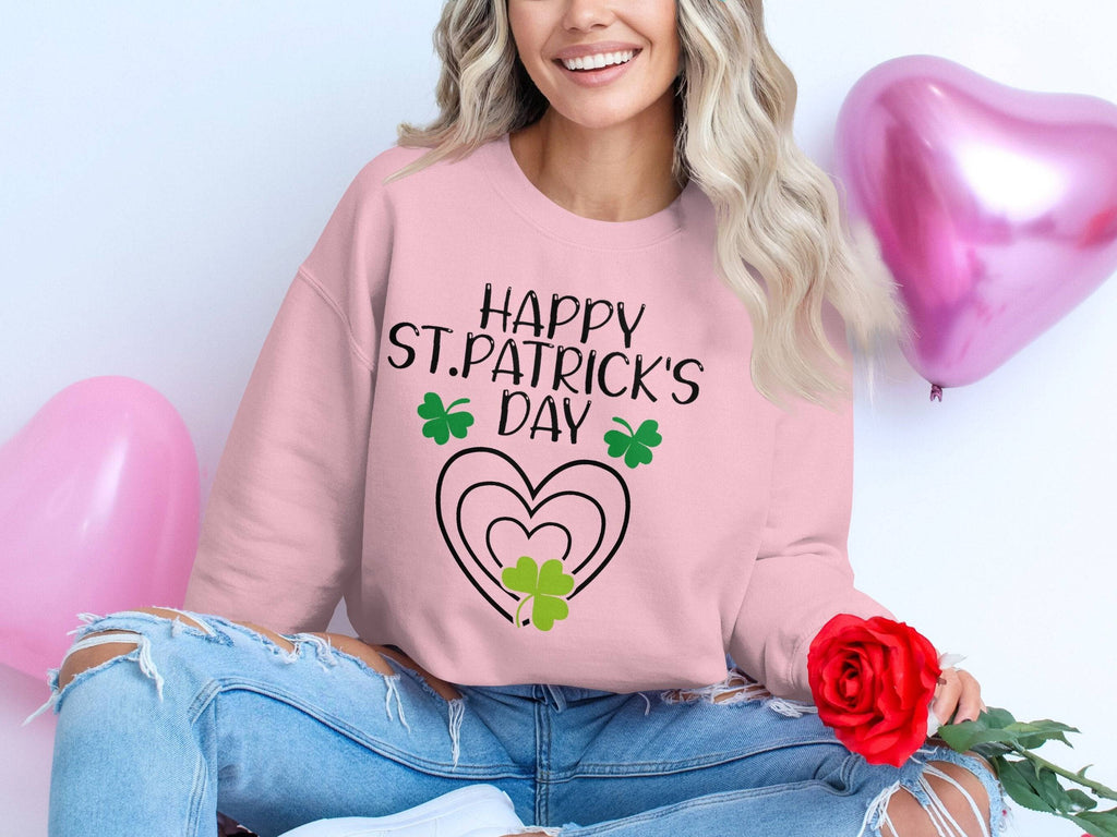 Wearing Garment Graphics festive sweatshirt adorned with Happy St. Patricks Day text and clover designs, a person with long wavy hair and ripped jeans sits cross-legged, holding a red rose amidst pink heart balloons.