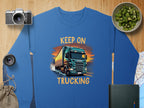 Garment Graphics offers a blue graphic sweatshirt featuring a truck with Keep On Trucking text, ideal for truck enthusiasts. Its surrounded by a camera, lens, kiwi bowl, book, cloth bag, and folded map on the wooden surface.