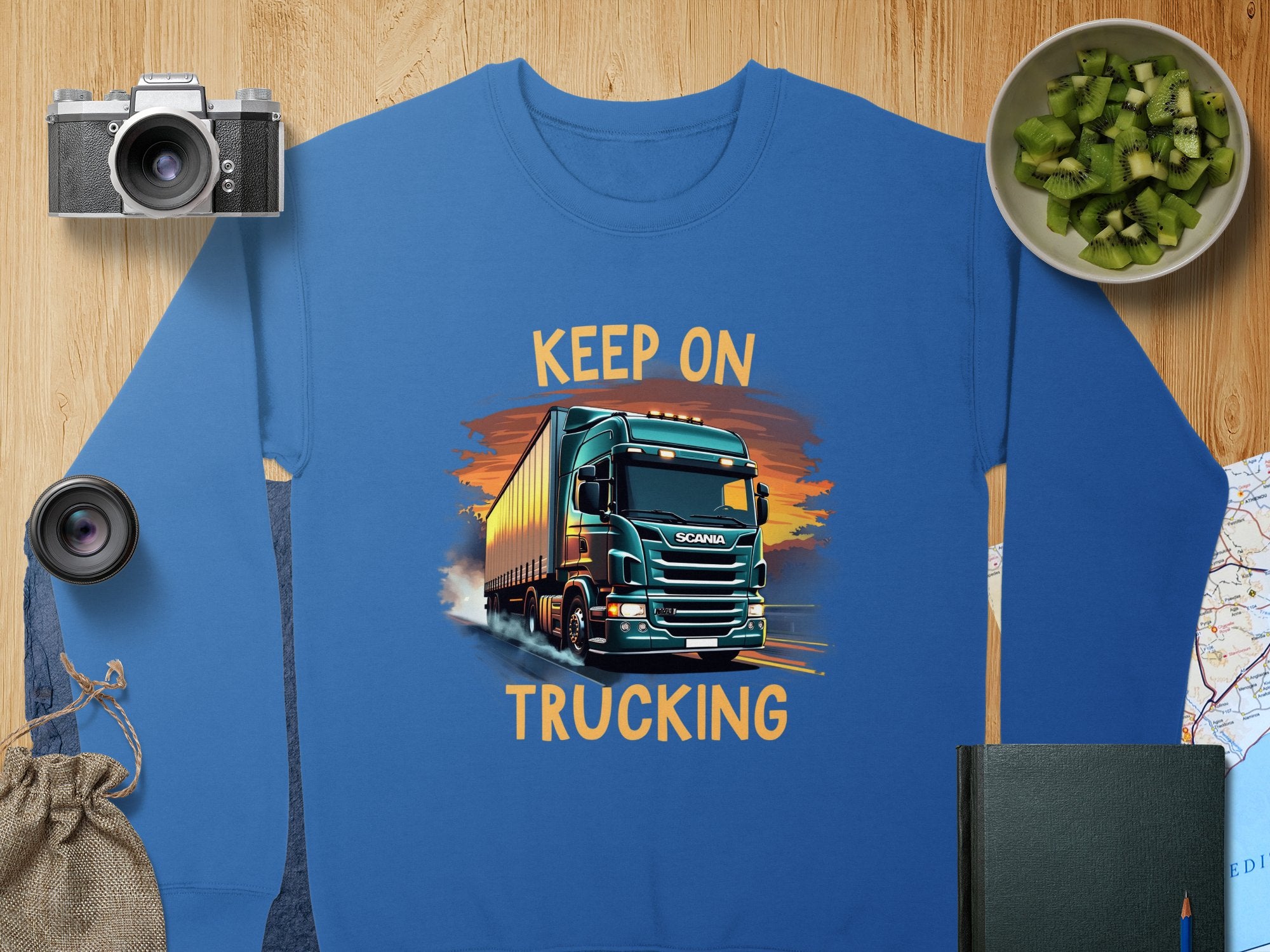 Garment Graphics offers a blue graphic sweatshirt featuring a truck with Keep On Trucking text, ideal for truck enthusiasts. Its surrounded by a camera, lens, kiwi bowl, book, cloth bag, and folded map on the wooden surface.