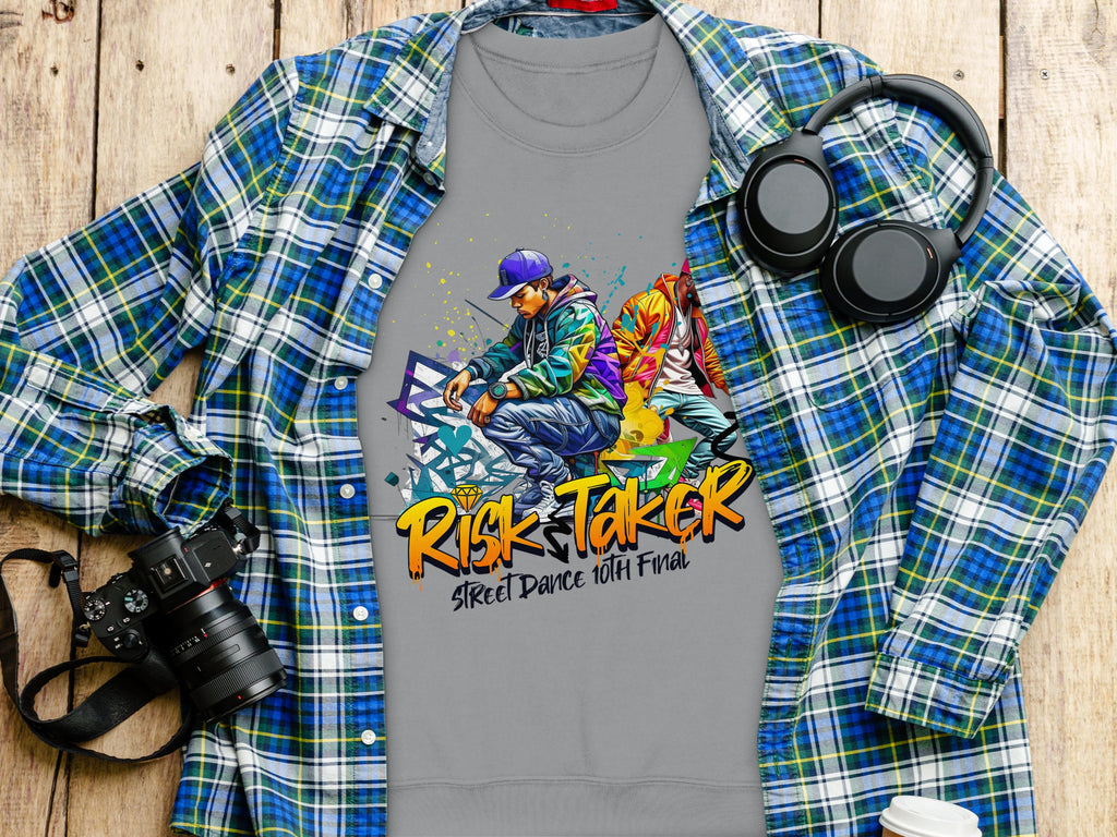 A gray Garment Graphics t-shirt with bold Risk Taker Street Dance 10th Final graphics is laid over a plaid shirt. A camera, headphones, and coffee cup nearby complete the effortless urban vibe on the wooden surface.