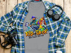 A gray Garment Graphics t-shirt with bold Risk Taker Street Dance 10th Final graphics is laid over a plaid shirt. A camera, headphones, and coffee cup nearby complete the effortless urban vibe on the wooden surface.