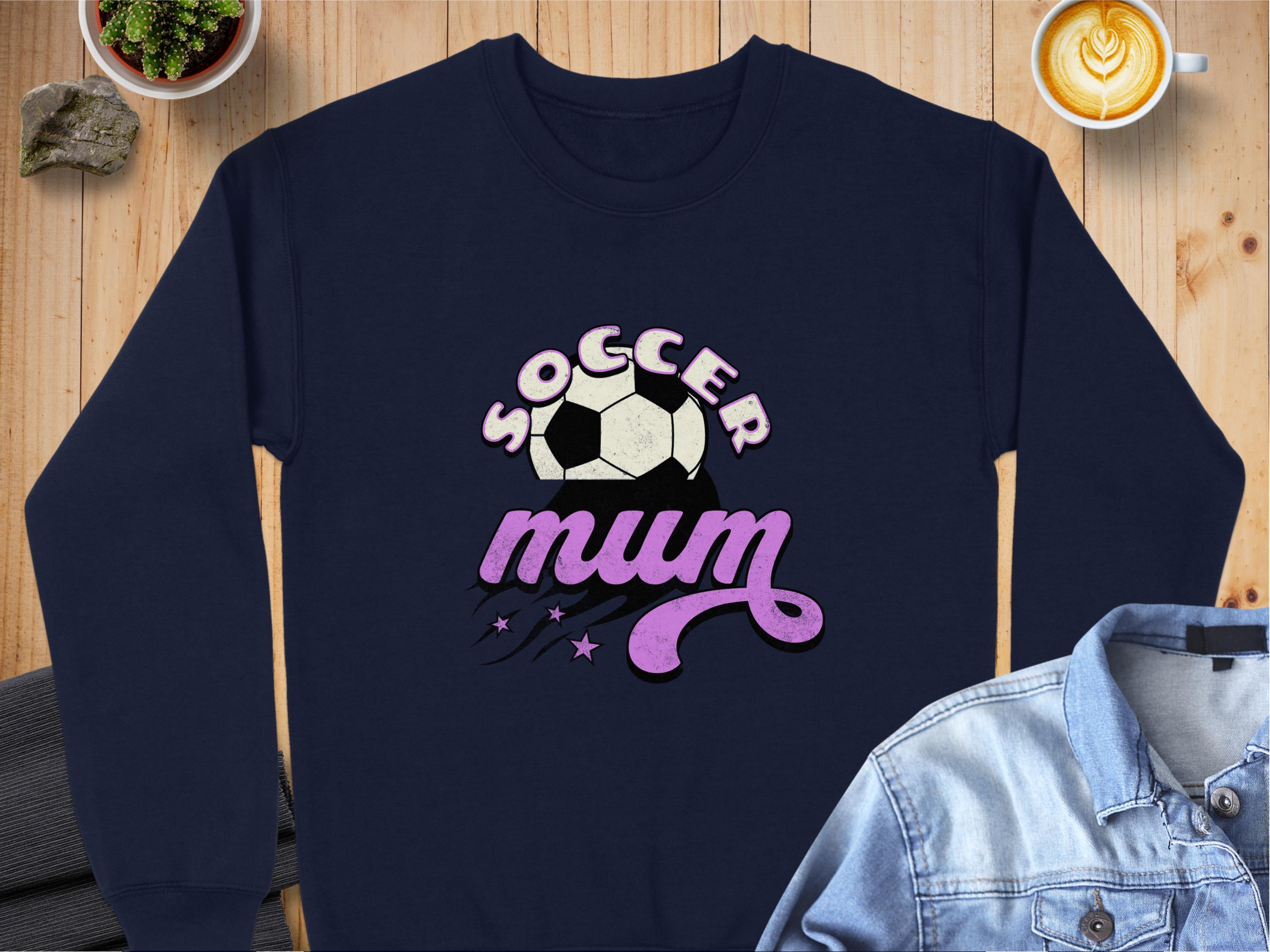 A navy sweatshirt from Garment Graphics, featuring Soccer Mum in vintage style with a soccer ball and pink lettering. Styled on a wooden surface with a denim jacket, potted plant, and latte for a cozy vibe.