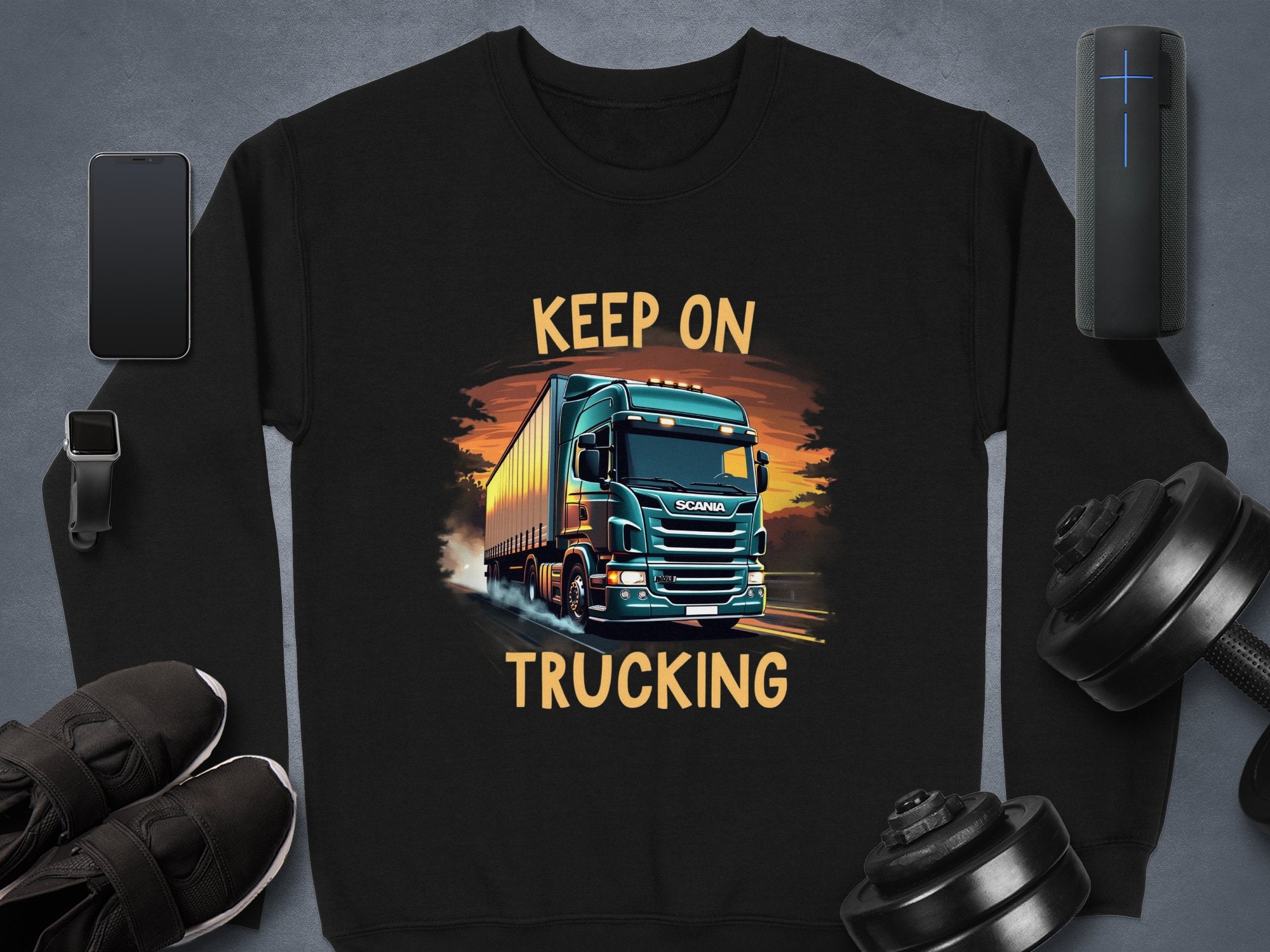 A Garment Graphics black sweatshirt features a bold truck design with orange and yellow hues, labeled Keep On Trucking. Accessories in the scene include a phone, smartwatch, shoes, speaker, and weights.