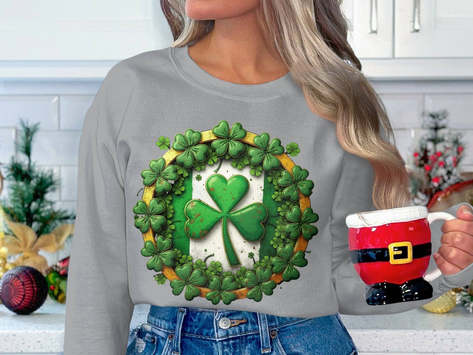 A person with long hair holds a Santa Claus-themed mug, embracing the Irish spirit. They wear a Garment Graphics gray sweatshirt with a large green shamrock surrounded by smaller ones resembling lucky four-leaf clovers. The background showcases white kitchen cabinets and festive holiday decor.