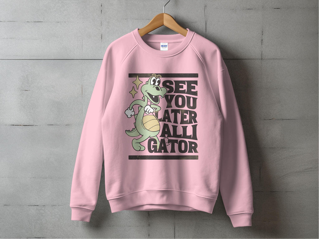 A pink sweatshirt from Garment Graphics hangs on a gray wall, featuring a cartoon alligator with a beach ball and stars. The text SEE YOU LATER ALLIGATOR is boldly displayed. This playful graphic sweatshirt comes in unisex sizes.