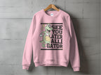 A pink sweatshirt from Garment Graphics hangs on a gray wall, featuring a cartoon alligator with a beach ball and stars. The text SEE YOU LATER ALLIGATOR is boldly displayed. This playful graphic sweatshirt comes in unisex sizes.