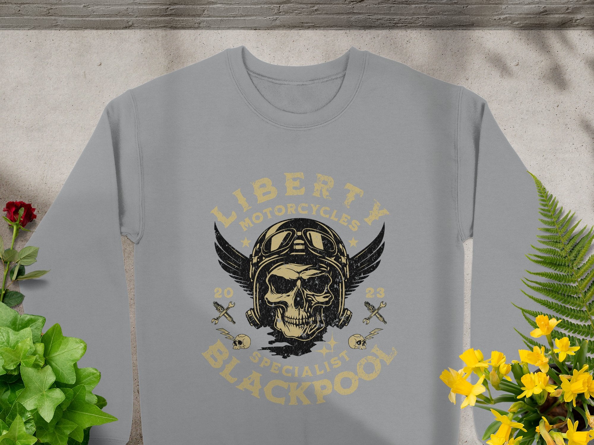 Garment Graphics presents a gray biker-style sweatshirt featuring a skull with goggles, a winged helmet, and the text Liberty Motorcycles Specialist Blackpool 2023 adorned with wrenches and stars. Surrounded by green plants and yellow flowers, it captures Liberty Motorcycles bold spirit.