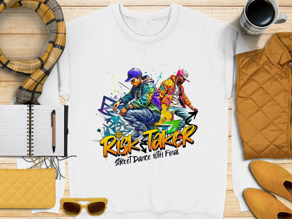 A white Garment Graphics sweatshirt with a vibrant graphic of two people dancing and the text Risk Taker Street Dance 10th Final lies on a wooden surface with notebooks, a pen, scarf, sunglasses, wallet, coffee cup, and yellow jacket—ideal for street dance finals.