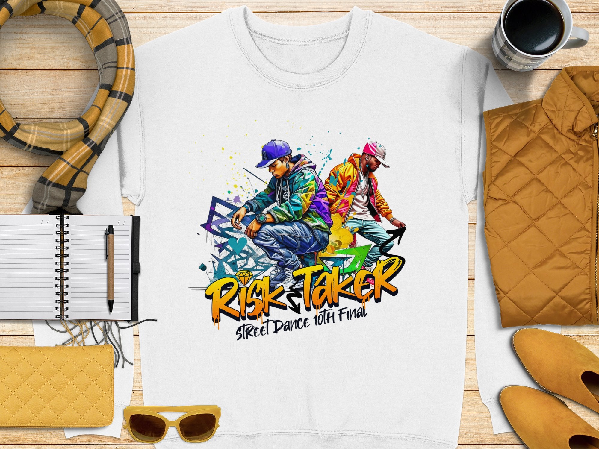 A white Garment Graphics sweatshirt with a vibrant graphic of two people dancing and the text Risk Taker Street Dance 10th Final lies on a wooden surface with notebooks, a pen, scarf, sunglasses, wallet, coffee cup, and yellow jacket—ideal for street dance finals.