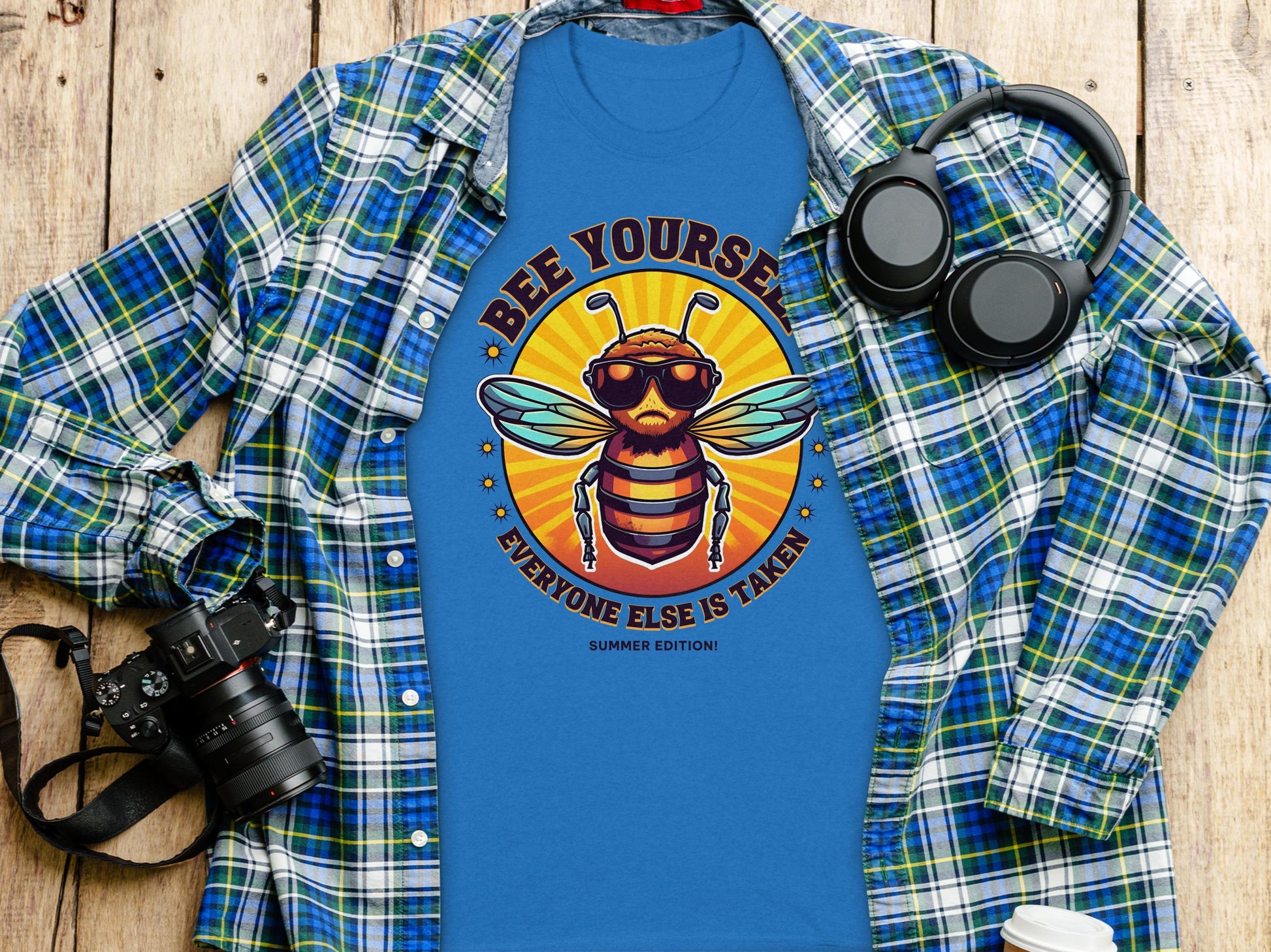 Blue T-shirt from Garment Graphics with a cool bee in sunglasses and headphones, surrounded by Bee Yourself, Every One Else Is Taken, Summer Edition! Placed on a plaid shirt with headphones, camera, and white tumbler on wood—a fun family collection addition.