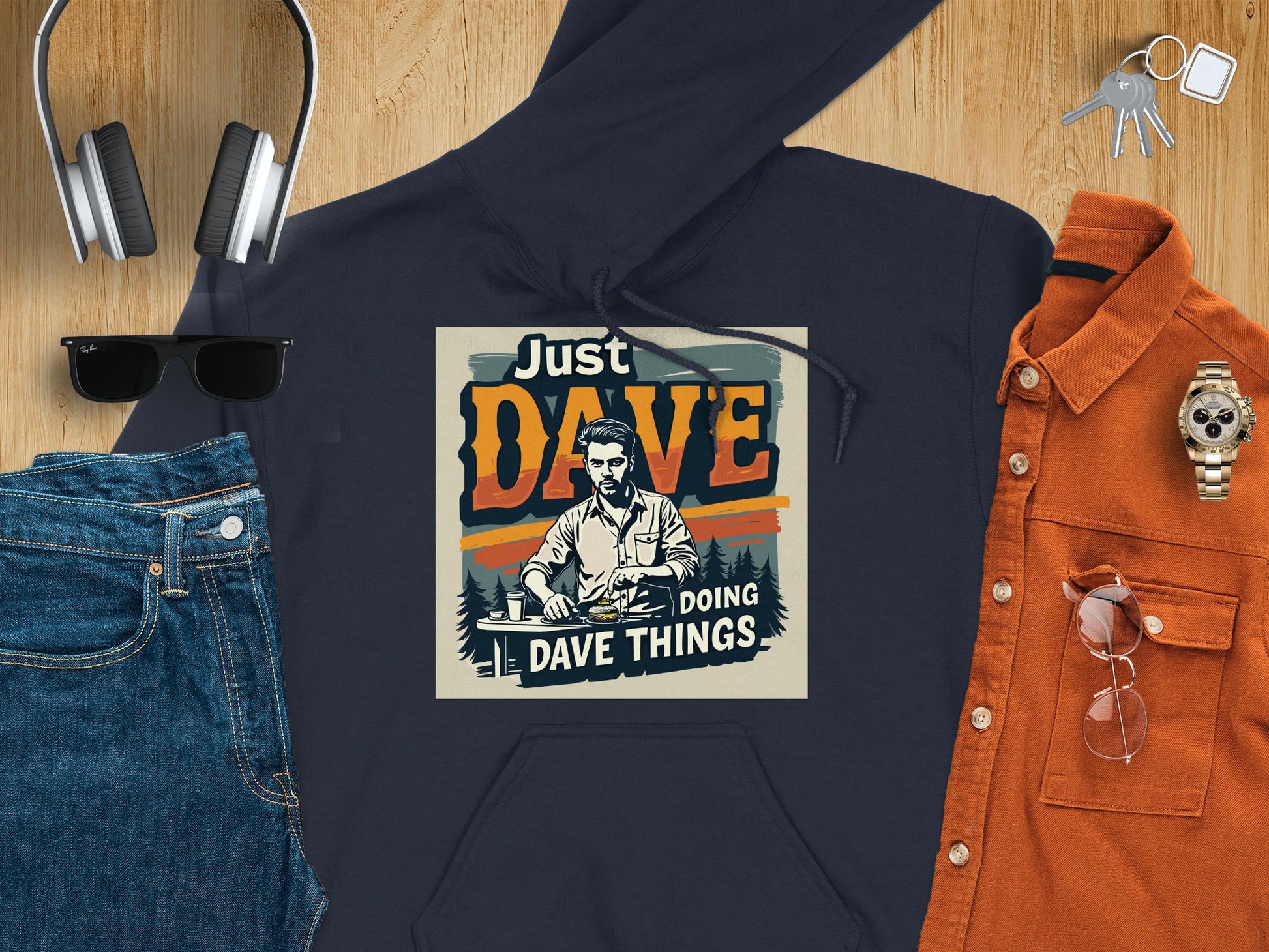 Flat lay showcasing Garment Graphics medium-heavy graphic hoodie with Just Dave Doing Dave Things design, teamed with blue jeans, an orange shirt, sunglasses, headphones, keys, and a watch on a wooden surface.