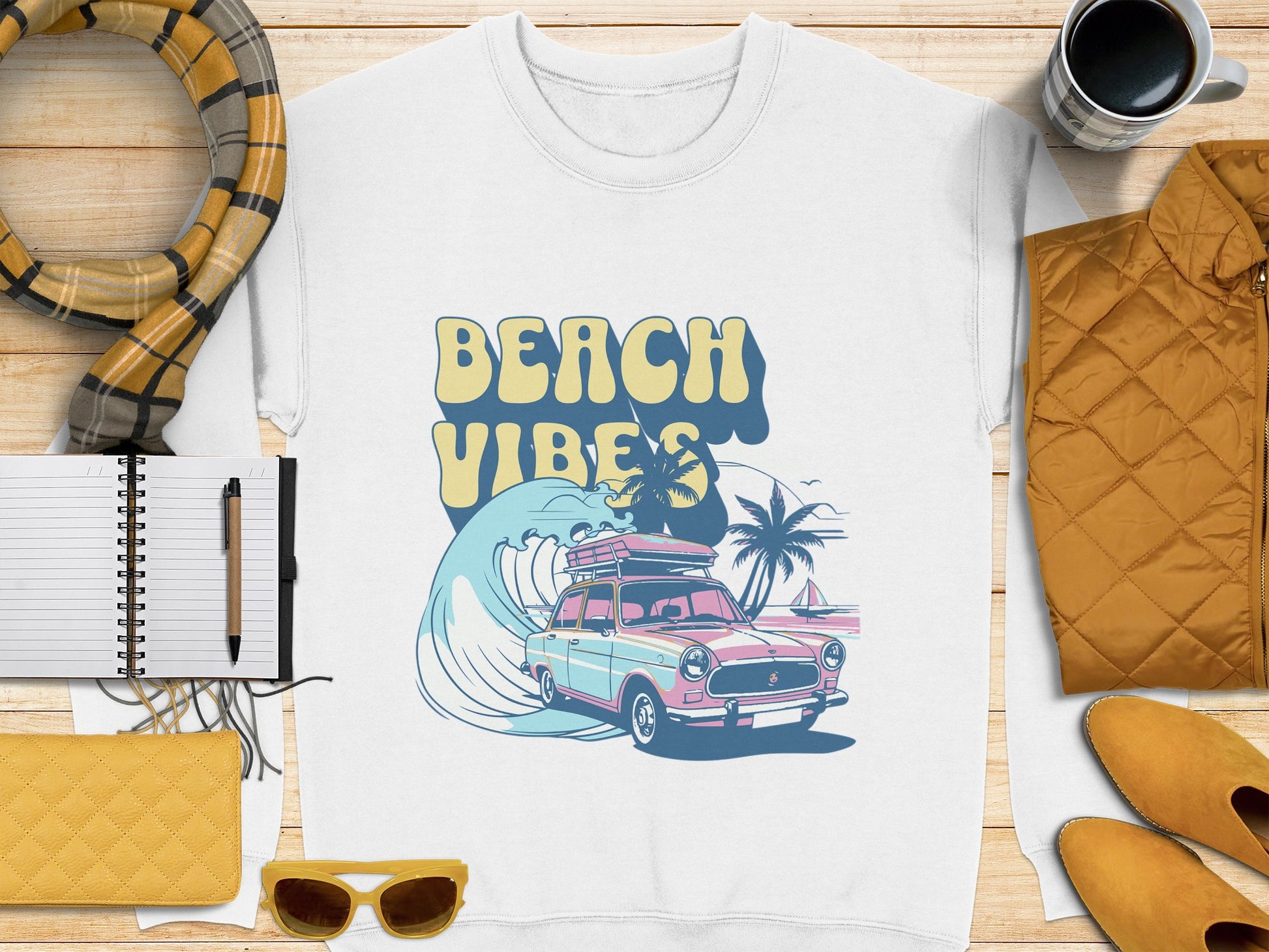 Flat lay of a white Garment Graphics T-shirt with a classic car, palm trees, and Beach Vibes text. It’s surrounded by a scarf, notebook, pen, yellow clutch, sunglasses, patterned jacket, and coffee mug on wood—all exuding retro vibes.