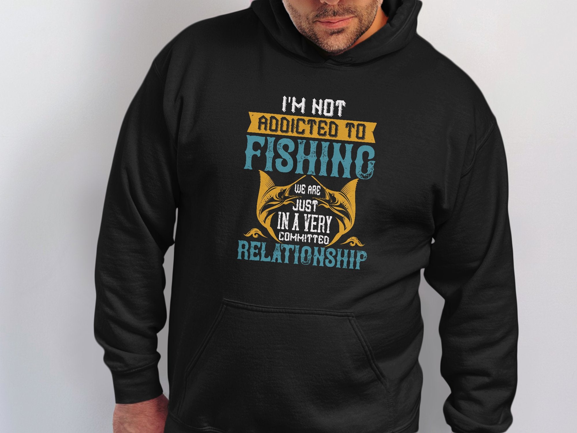 A person wears a Garment Graphics black fishing hoodie, featuring the humorous design, Im not addicted to fishing. We are just in a very committed relationship. This classic fit piece seamlessly combines comfort and style for dedicated anglers.