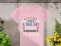 A pink Garment Graphics womens fishing T-shirt with the text Id rather have a bad day fishing than a good day at work is showcased on concrete, surrounded by vibrant green plants and yellow flowers, ideal for fishing enthusiasts.